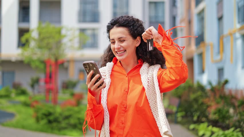 Excited woman in orange shirt and white shawl makes online purchases using phone and bank card. She feels happy and enjoys shopping in courtyard.