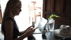 A mature woman sits at a cafe table engrossed in her tablet and phone while enjoying a coffee. She embodies the digital nomad lifestyle in a warm environment. - Powered by Shutterstock - Get 15% off with code: PIKWIZARD15