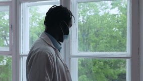 Exhausted african american healthcare professional removing protective mask, gazing pensively through hospital window after intense pandemic shift or patient loss. Sad doctor surgeon looking  - Powered by Shutterstock - Get 15% off with code: PIKWIZARD15