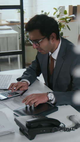 Vertical shot of busy Indian CEO man working with documents and typing on laptop at desk in modern bank office