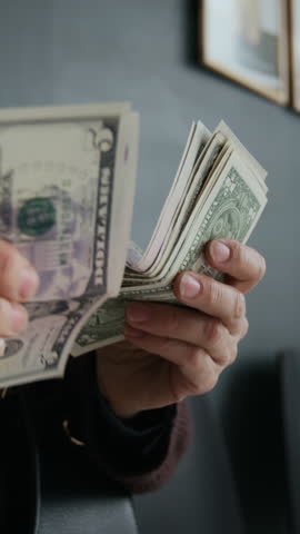 Vertical close-up shot of unrecognizable hands of elderly woman counting stack of American dollars in bank office