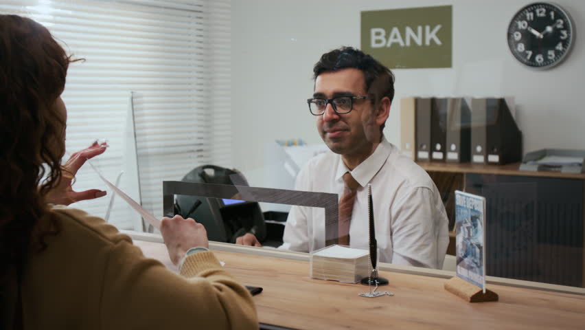 Over the shoulder shot of unrecognizable woman with red curly hair interacting with bank assistant through glass teller window in office