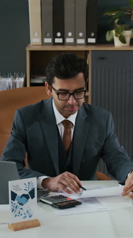 Vertical shot of Indian businessman sitting at cluttered desk and hard-working in his modern office