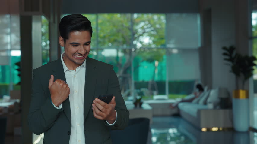 Portrait of joyful Indian man in formal suit uses smartphone stock market app indoors. Young man entrepreneur celebrates job promotion via email standing in company office