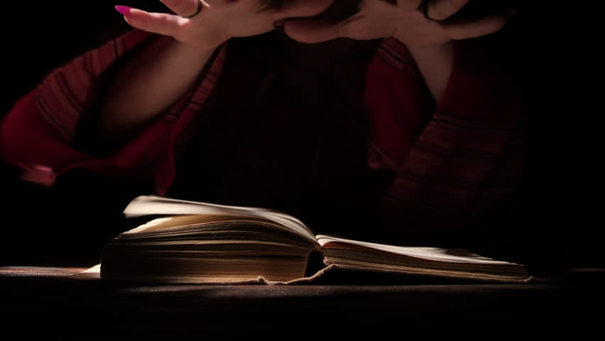 Witch hands casting spells over magic book in dark room close up