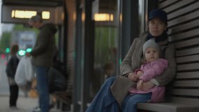 Passengers at a public transport stop. Mother and little daughter sitting on bus stop bench, patiently waiting for a bus and hugging. Evening time of day. Transportation and traffic concept video. - Powered by Shutterstock - Get 15% off with code: PIKWIZARD15