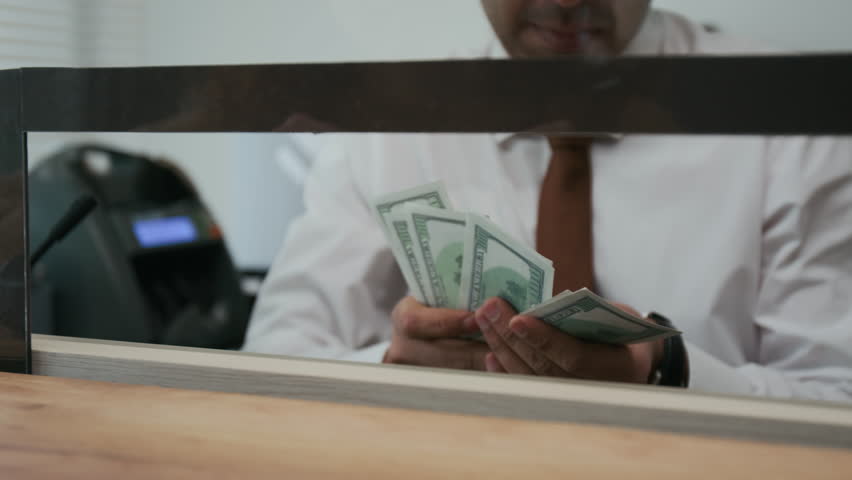Close-up low angle shot of male Indian bank teller, wearing white shirt and tie, counting stack of American dollar bills