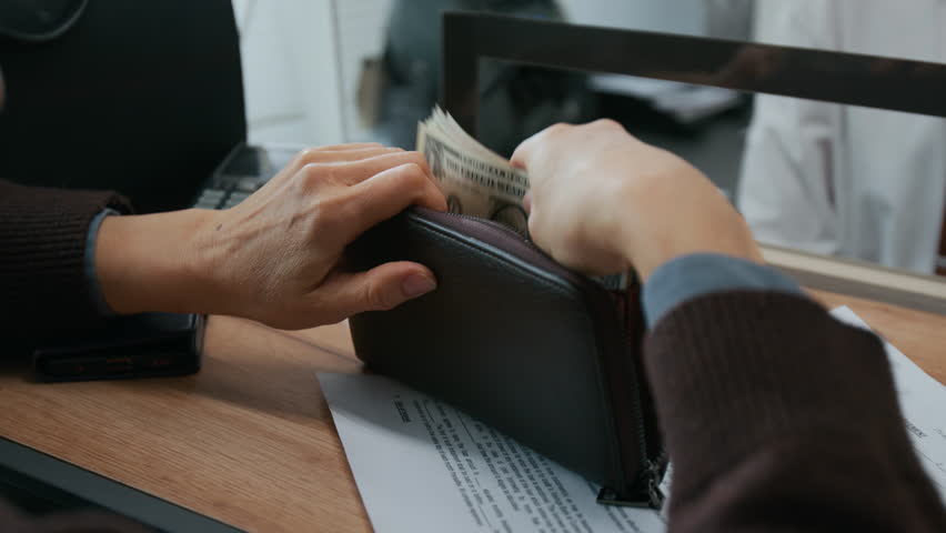 Close-up shot of unrecognizable elderly woman giving stack of dollar bills to male bank worker through glass teller window
