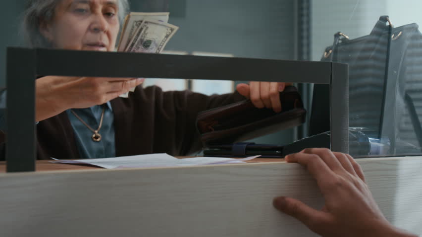 Low angle shot of unrecognizable bank worker counting dollar bills given by elderly customer through glass teller window