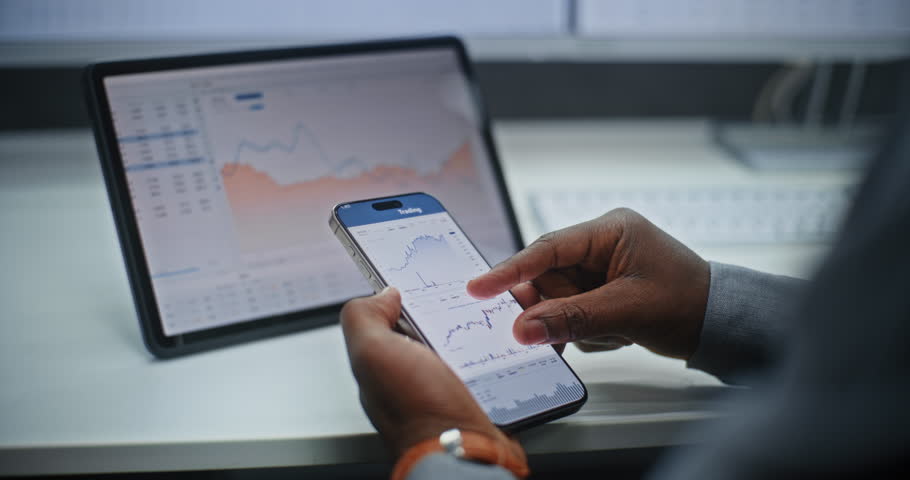 African American Stock Exchange Broker Analyzing Real-Time Stocks and Exchange Market Charts on Smartphone and Digital Tablet Using Online Trading App. Investment Agent Working in Office. Close Up.