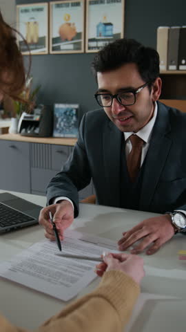 Vertical shot of businessman in suit showing where to sign document to unrecognizable woman at desk in his office