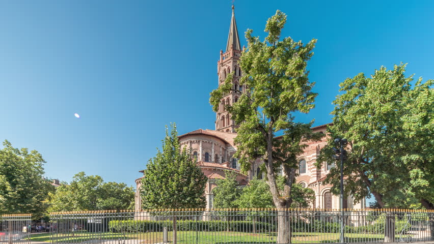 The cobbled parvis of the Basilica of Saint Sernin in Toulouse, France, timelapse hyperlapse. Largest romanesque church in Europe with brick bell tower, arched doors, green trees and blue sky