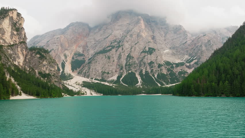 Aerial shot flying just above Lake Braies, reflecting the cloudy sky and slowly unveiling the majestic Dolomite range in the background during a quiet spring day