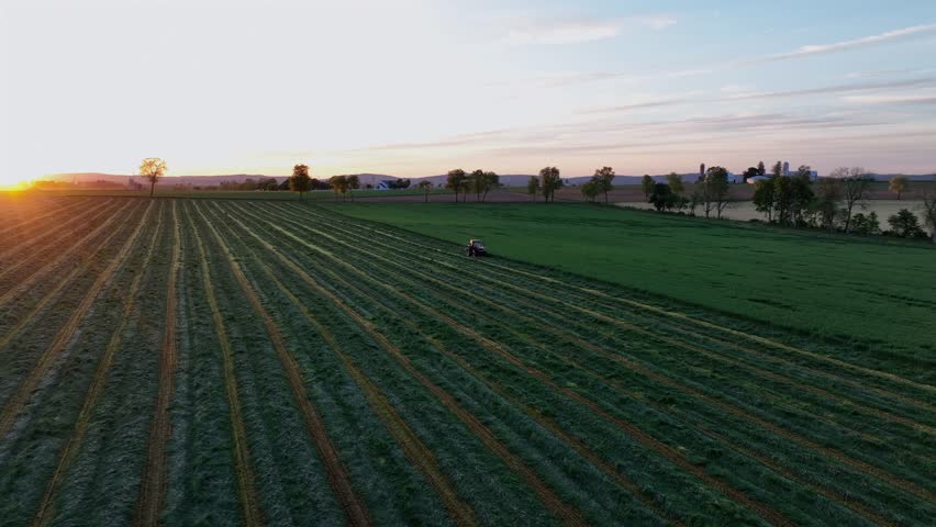 Tractor with machinery working on farm field in american countryside at sunrise. Aerial orbit shot. Rural farm fields in May. Wide shot.