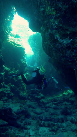 Vertical footage, Group of scuba divers as they swims inside crevice between coral reef in sun rays, Slow motion, Adventure and marine exploration concept, scubadivers exploring deeps of Ocean