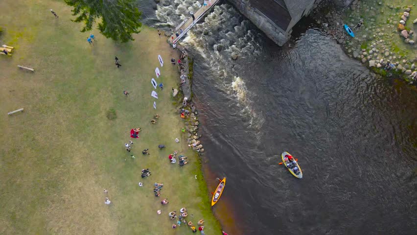 Top down aerial view of SUP mega paddleboards and colorful kayaks going through foamy river rapids on a narrow river from underneath a bridge during Võhandu marathond while spectators watch on sides.
