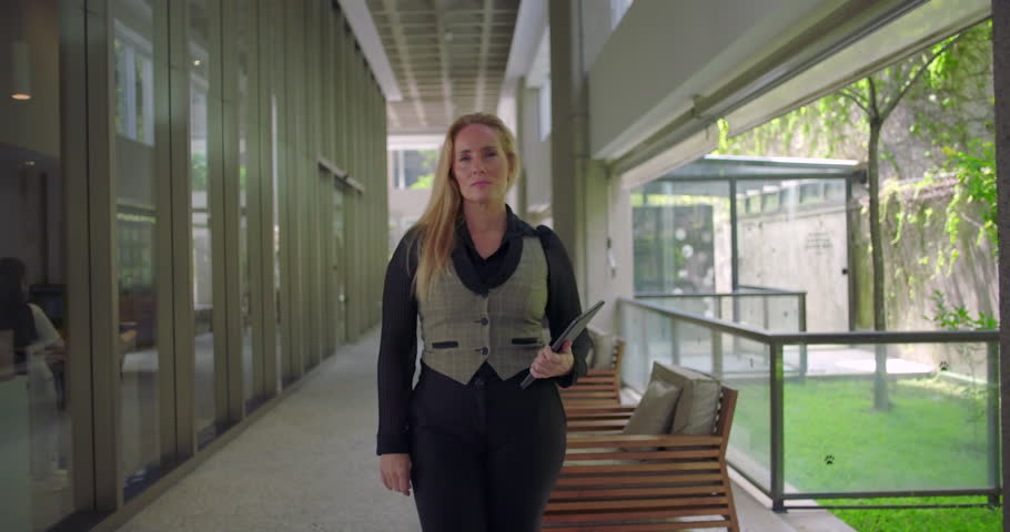 Woman standing still in modern hallway holding tablet, facing camera with neutral expression, prepared for professional task or meeting