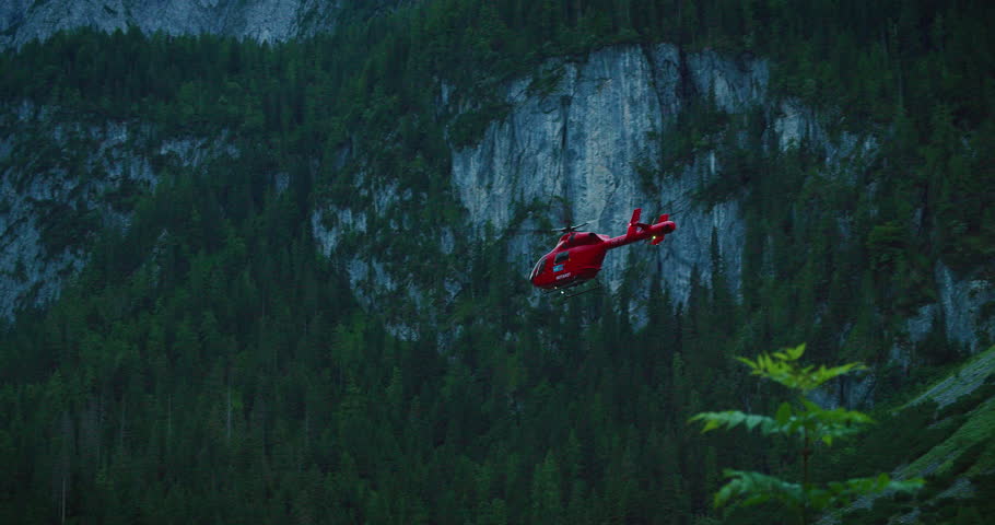 Red rescue helicopter flying over lush green Austrian mountain landscape with rocky cliffs