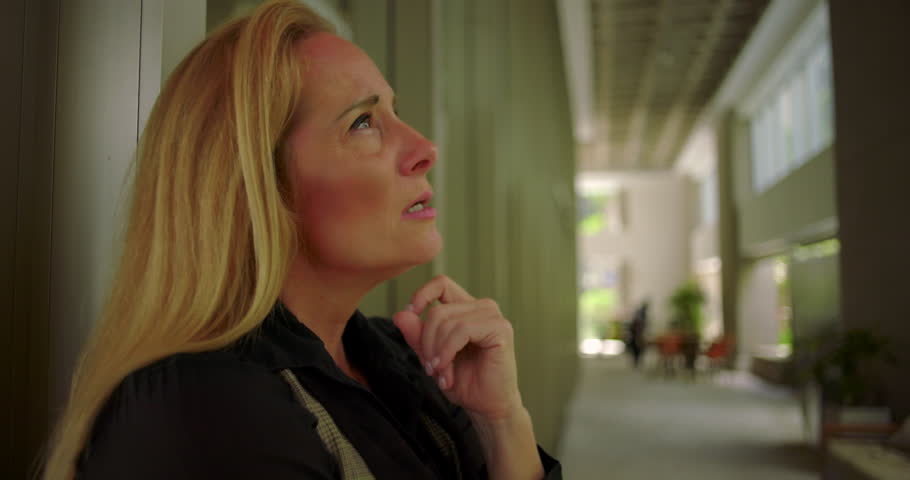 Side view of woman lowering her gaze with hand near chest in corporate corridor with modern seating and soft natural light through windows