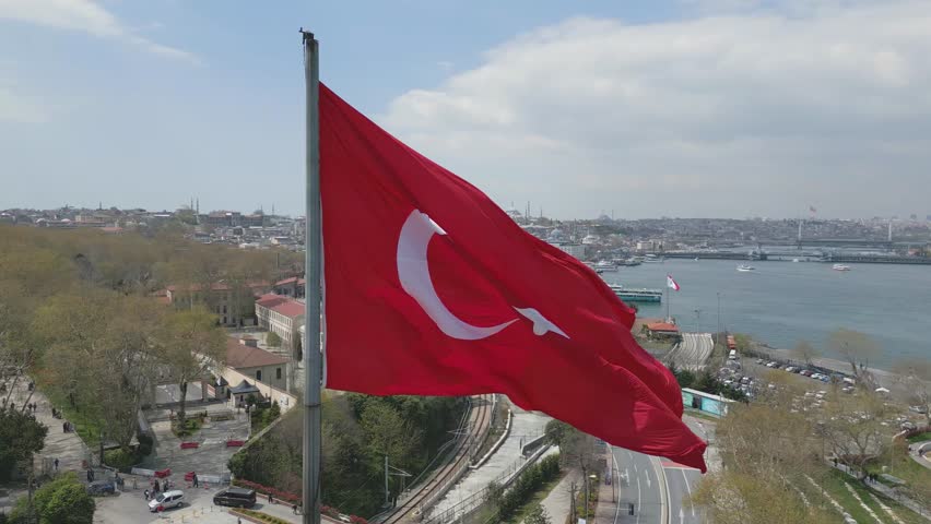 A vibrant Turkish flag proudly flutters in the wind, overlooking the Bosphorus Strait. Istanbul's bustling skyline contrasts beautifully with the serene waters below.