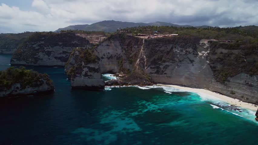 This aerial view of Diamond Beach in Bali reveals a stunning coastline under sunny skies where turquoise ocean meets white cliffs and tropical waves crash against the remote shoreline.