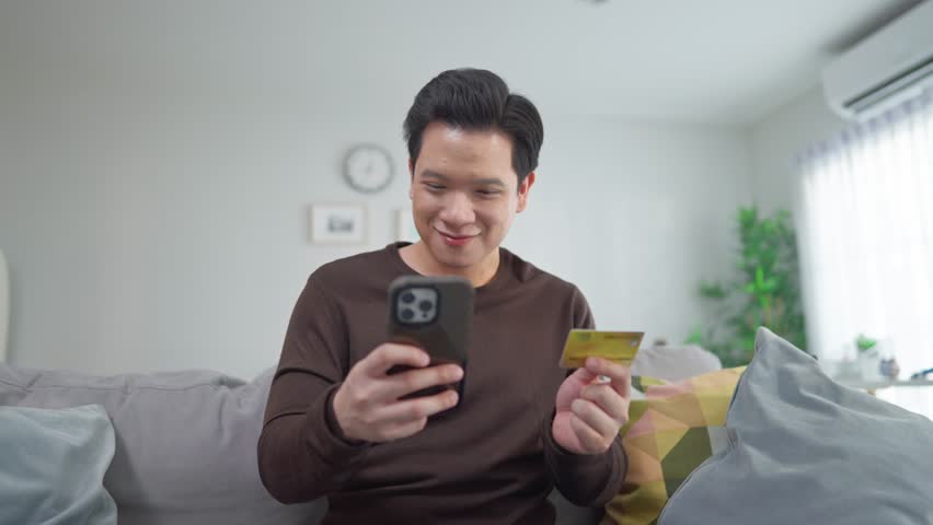 Asian man using mobile phone shopping online in living room at home. Attractive mature man sit on sofa, holding credit card make payment after use smartphone purchase goods in web store in house.