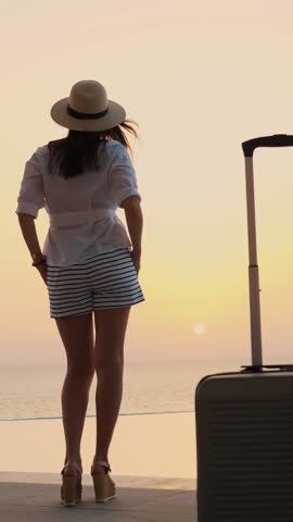 Back view of young woman in summer clothes and sun hat, with travel luggage, standing at the pool, enjoying the sunrise. summer. seaside resort. travel concept.