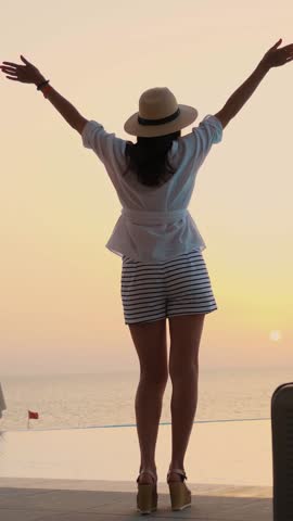 Back view of young woman in summer clothes and sun hat, with travel luggage, standing at the pool, enjoying the sunrise. summer. seaside resort. travel concept.