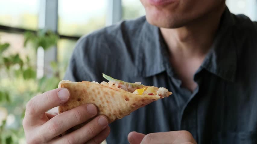 A man in a gray shirt sits at a table in a restaurant, enjoying a slice of pizza. Ideal 4K video for food ads, restaurant promos, and casual dining content.