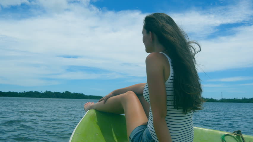 Happy woman sits on bow of boat and admires a beautiful nature landscape during trip. Pretty girl with blowing long hair rests on deck of yacht and enjoys summer travel. Vacation or holiday concept