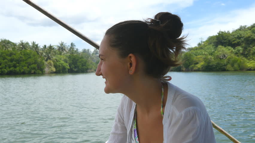 Young woman with blowing long hair sits on bow of boat and looks at beautiful nature landscape. Unrecognizable girl relaxes on deck of yacht and enjoys summer travel. Concept of vacation or holiday