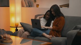 Multiracial lesbian couple enjoying cozy evening on sofa, cuddling and smiling while watching laptop together, sharing an intimate moment in warm, relaxed home atmosphere. High quality 4k footage - Powered by Shutterstock - Get 15% off with code: PIKWIZARD15