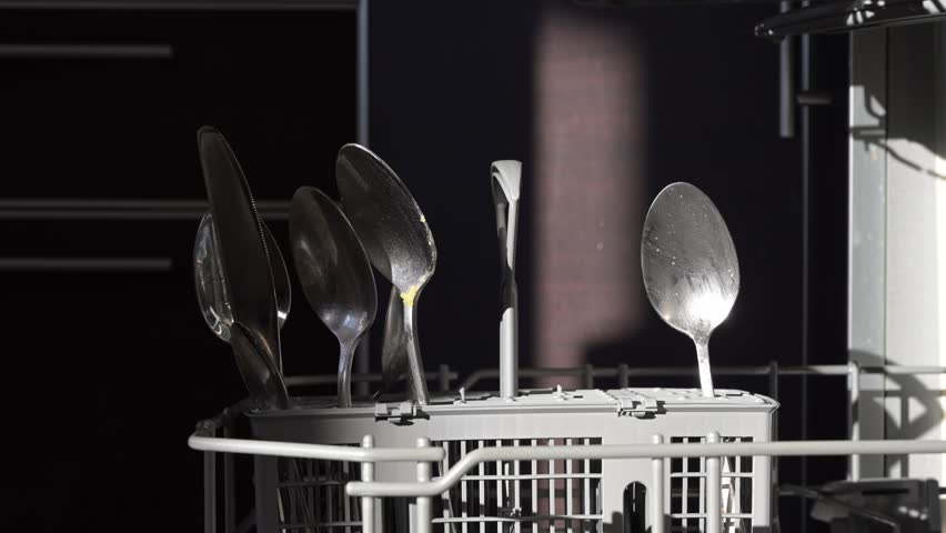 Stainless steel spoons gleaming brightly, sunlight casting reflective highlights across clean kitchen utensils drying in dishwasher basket