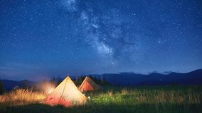 Blurred silhouettes of tourists resting at campsite beside two glowing tents on grassy hillside under star-filled sky with Milky Way. Beauty of clear night in nature, with mountains in background. - Powered by Shutterstock - Get 15% off with code: PIKWIZARD15
