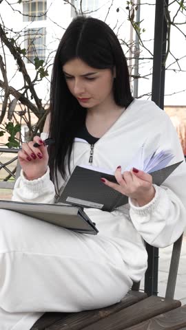 A female student sits on a park bench and studies using a tablet and takes notes.