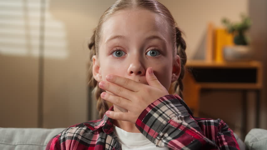 Portrait surprised little school girl child kid daughter preteen looking at camera at home sofa couch big eyes close mouth with hand feel stunned amazed unexpected news shocked unbelievable surprise