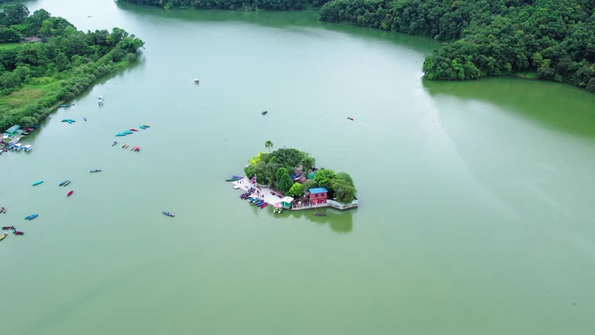 Aerial view of a small island in a lake, surrounded by boats and lush greenery, with a pathway and buildings on the shore. 