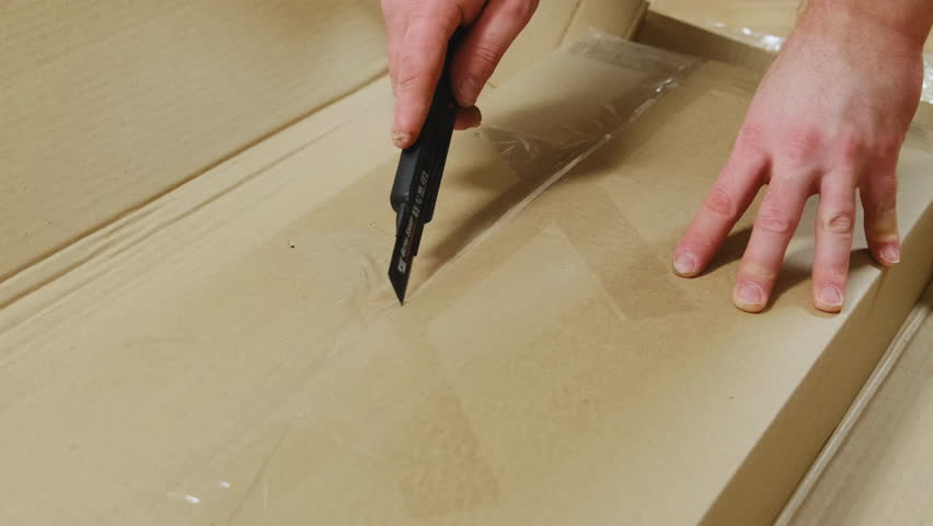Man with blade cuts adhesive scotch tape on cardboard box in room closeup. Handyman uses sharp knife to unpack carton package with supplies