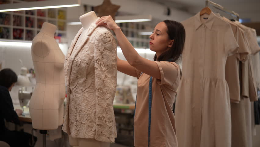 Dressmaker taking measurements of white lace jacket on mannequin, working in sewing workshop with other tailors and various clothes and supplies. Concept of sewing manufacturing, slow motion.