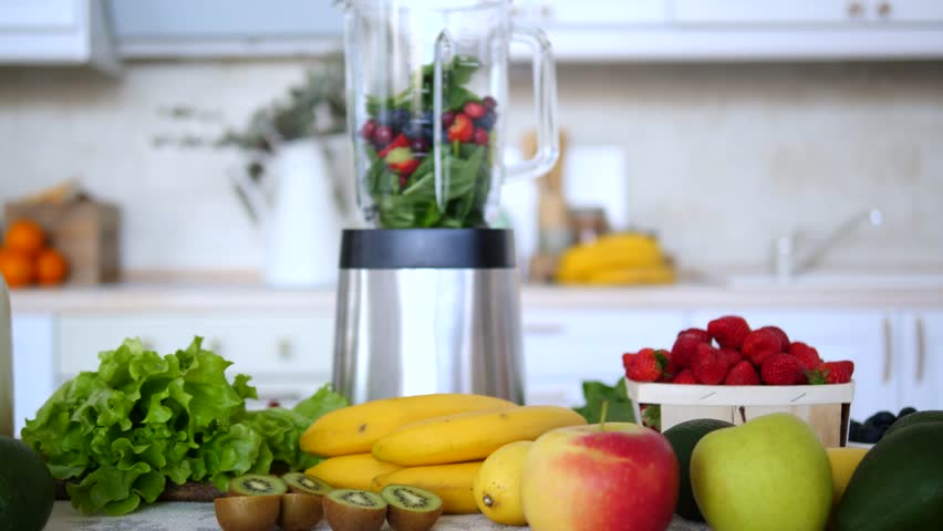 Fresh berries, spinach, and other fruits are placed in a blender, ready to be mixed into a healthy smoothie, surrounded by various fruits and vegetables on a kitchen counter