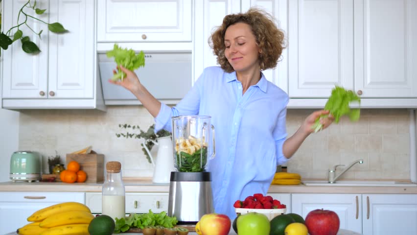 Cheerful woman dances with lettuce leaves in her hands, preparing a healthy green smoothie in her modern kitchen, surrounded by fresh fruits