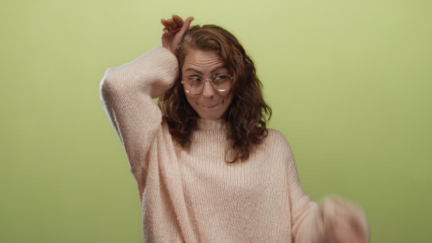 Young woman making playful gestures with hands on head, wearing glasses and sweater, standing against isolated yellow background, exuding a fun and cheerful mood.