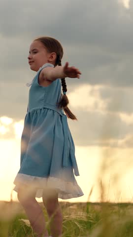 Child girl in dress dances spinning with her arms out to sides. Child dream to dance. Happy child girl plays spinning in park on grass. Little girl smiles in front of sky. Children's dreams in outdoor