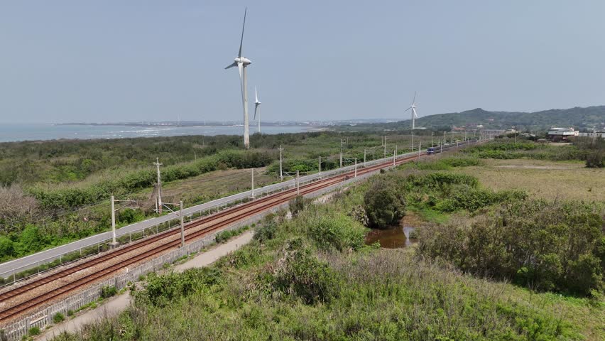 Aerial view of a regional train dashing through green fields on a sunny day and giant wind turbines standing by the beach in a seaside park on the windy coast, in Houlung, Miaoli County, Taiwan, Asia