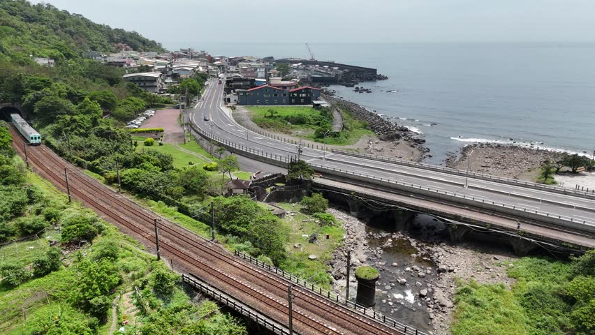 A tourist train (Sea Breeze) coming out of a tunnel and dashing thru a bridge over a little stream by the mountainside with Daxi Fishing Harbor in background in Toucheng Township, Yilan County, Taiwan