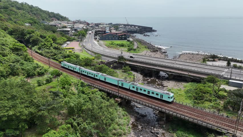 A tourist train (Sea Breeze) coming out of a tunnel and dashing thru a bridge over a little stream by the mountainside with Daxi Fishing Harbor in background in Toucheng Township, Yilan County, Taiwan
