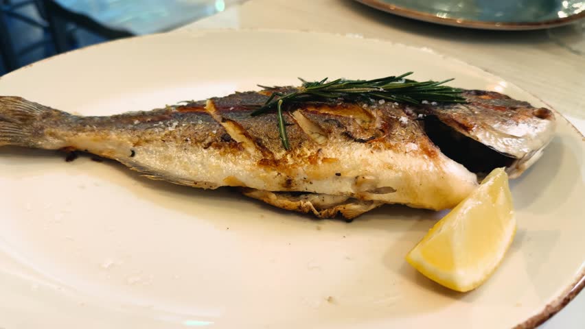 Grilled white fish on a plate served with a lemon wedge and a sprig of rosemary. High quality 4k footage