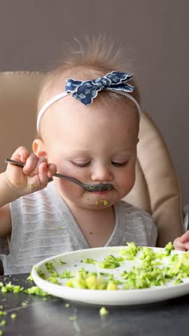 Messy baby girl eating healthy food broccoli with spoon at table, adorable toddler learning to feed herself, cute child mealtime chaos, candid moment of childhood