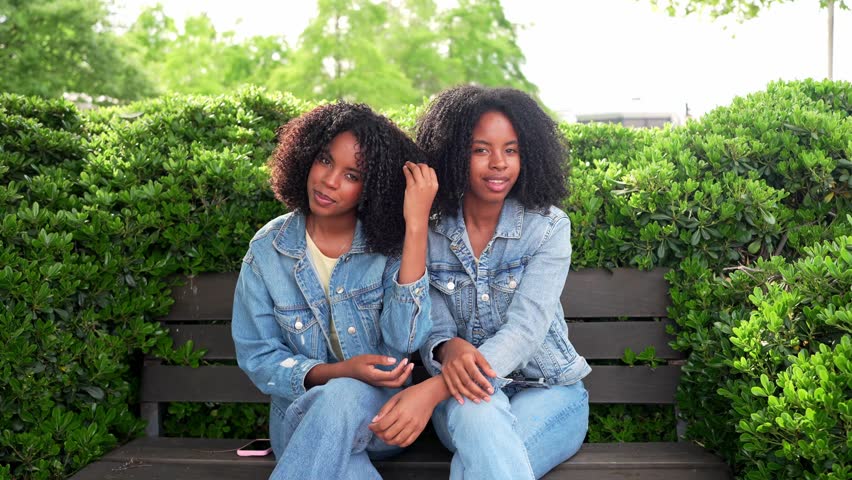 Two twin sisters wearing denim jackets are holding hands while sitting together on a park bench near a green hedge
