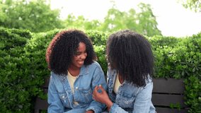 African two happy twin sisters are sitting on a park bench, wearing denim jackets and laughing together on a sunny day - Powered by Shutterstock - Get 15% off with code: PIKWIZARD15